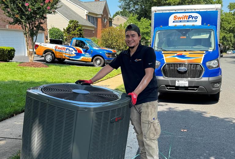 A smiling SwiftPro technician with his hands on a Trane air conditioning condenser on a Northern Virginia driveway, a wrapped SwiftPro box truck and pickup behind him